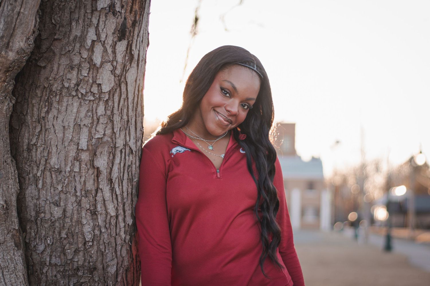 Smiling young woman in a red jacket leaning against a tree at golden hour outdoors.