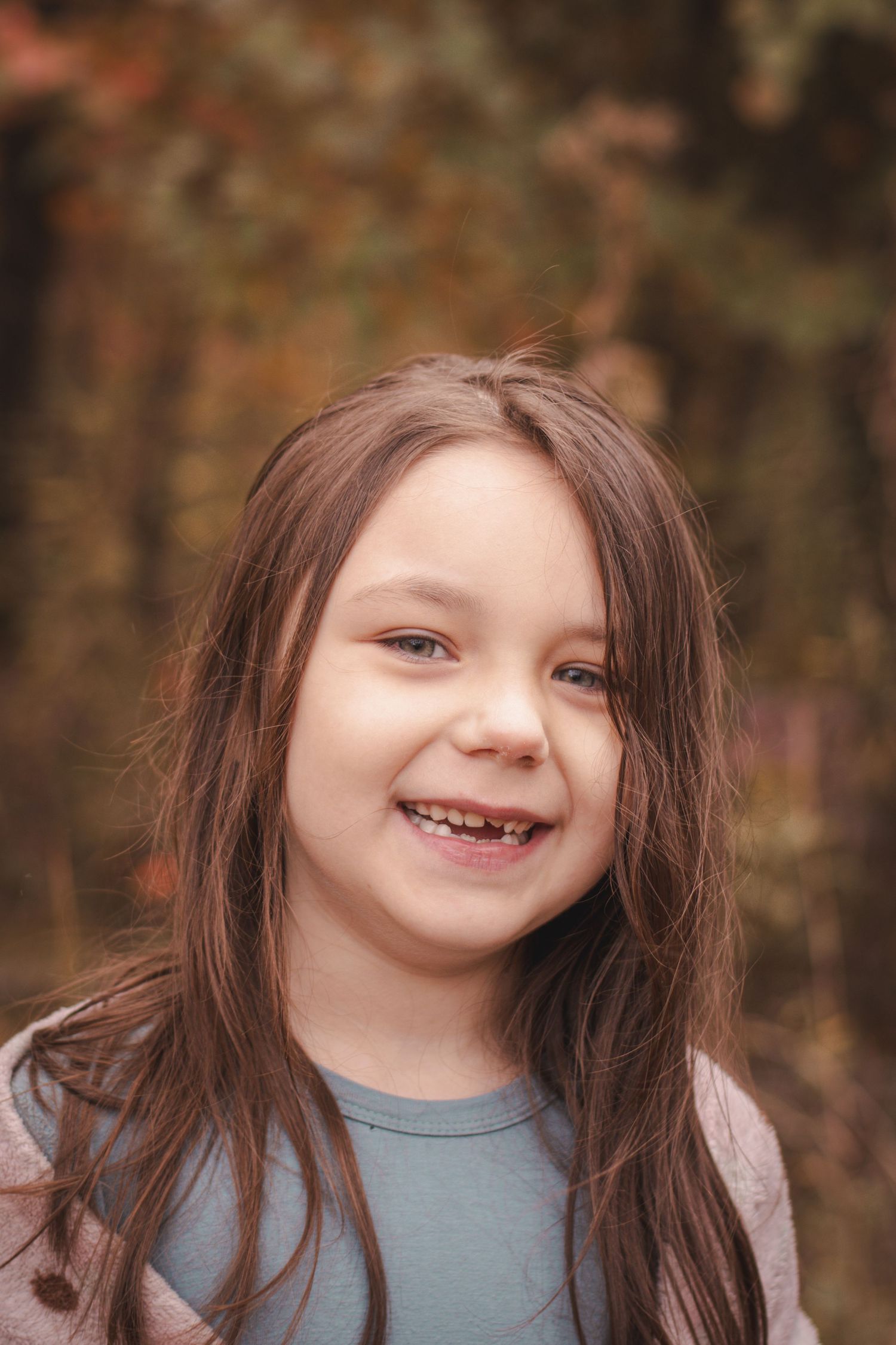 A smiling young girl with long brown hair and missing front teeth poses outdoors with fall foliage in the background.