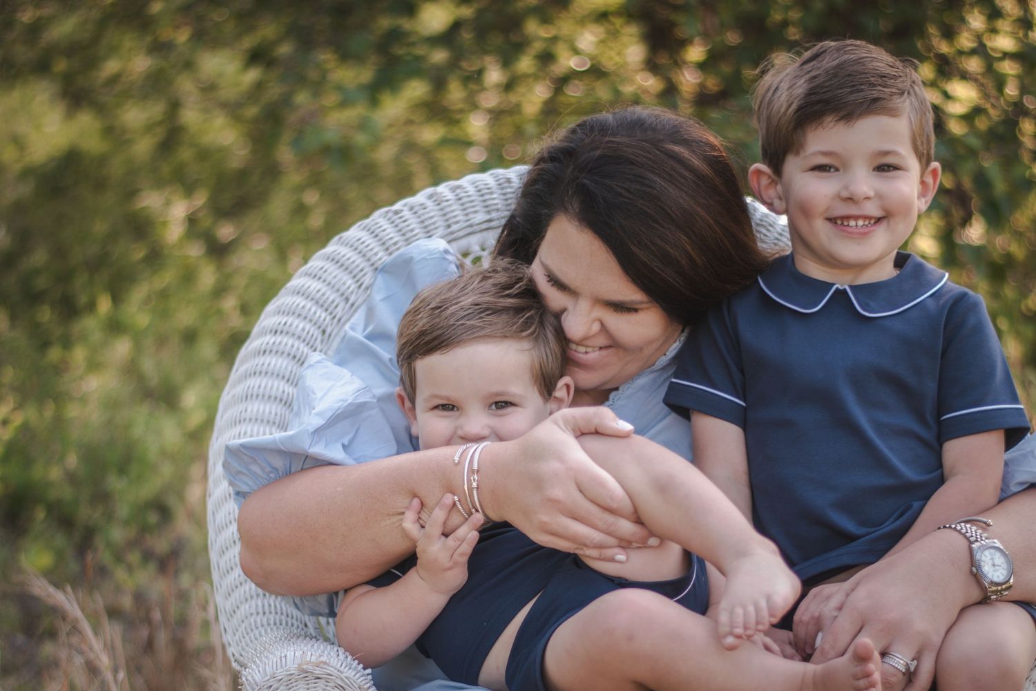 A mother hugs her two young boys in a wicker chair outdoors, smiling warmly in a lush green setting.