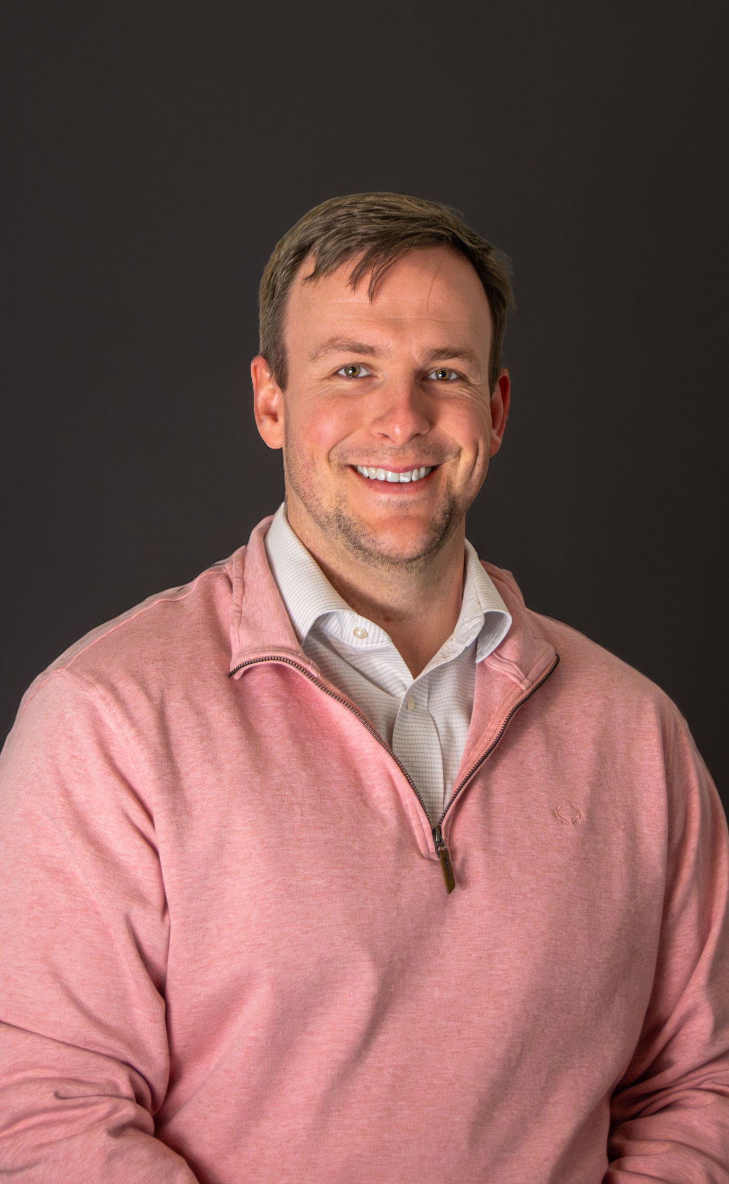 Smiling man in pink quarter-zip pullover poses for professional headshot against dark gray background.