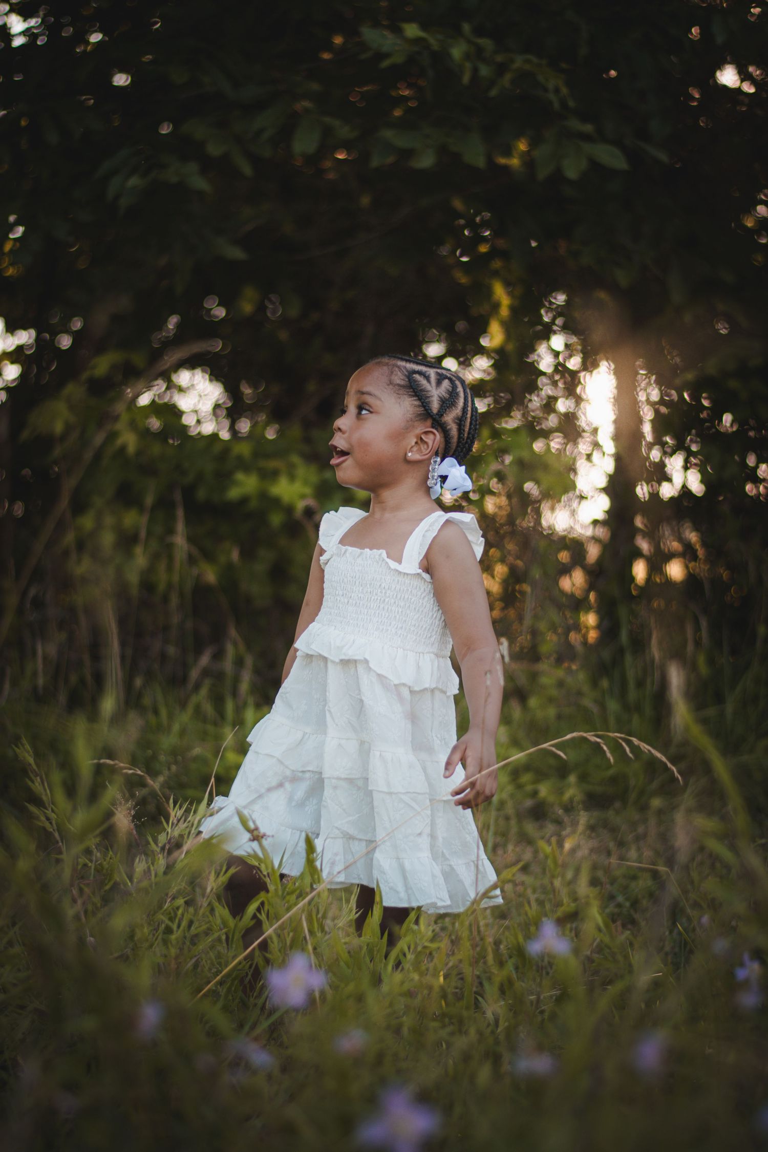 Young girl in white ruffled dress standing in lush green field at golden hour sunset.