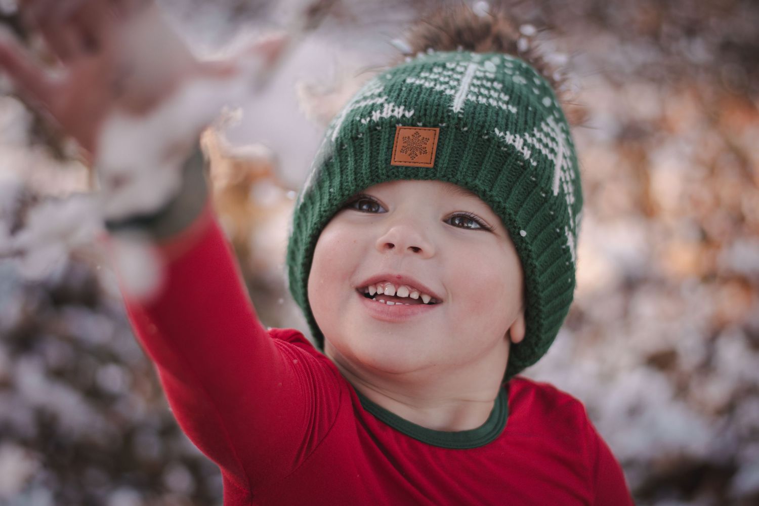 Happy young boy in a green winter hat and red shirt playing in the snow outdoors.