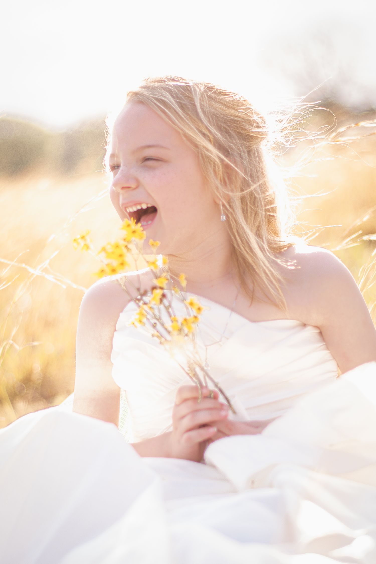 Joyful blonde bride laughing in sunlit golden field holding small yellow wildflowers.