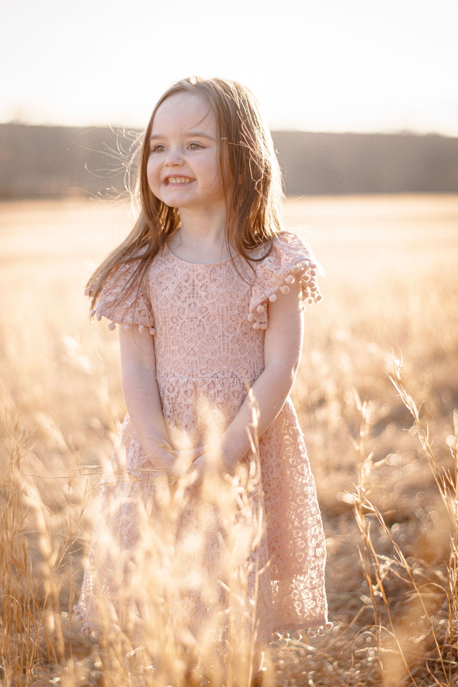 Young girl in pink lace dress smiling joyfully in a golden wheat field during sunset.