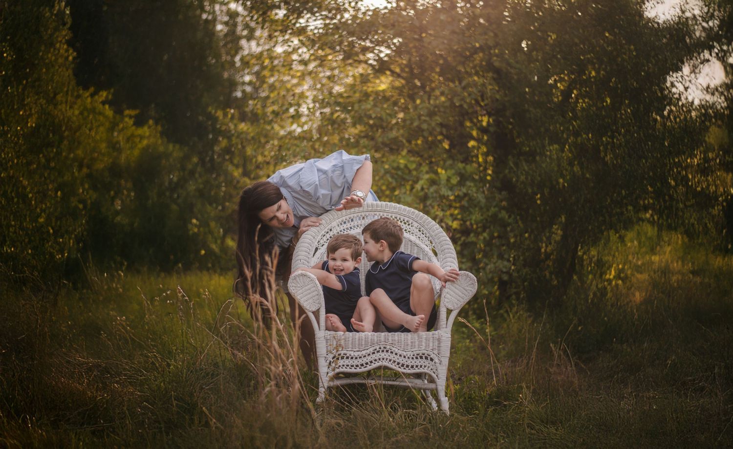 A mother leans over two children sitting in a white wicker chair surrounded by lush greenery in golden hour light.
