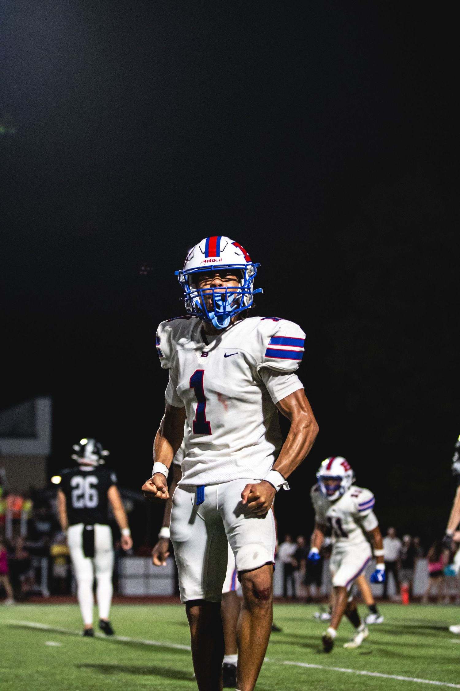 Football player wearing white and blue uniform with number 1 jersey and helmet walks on field during night game.