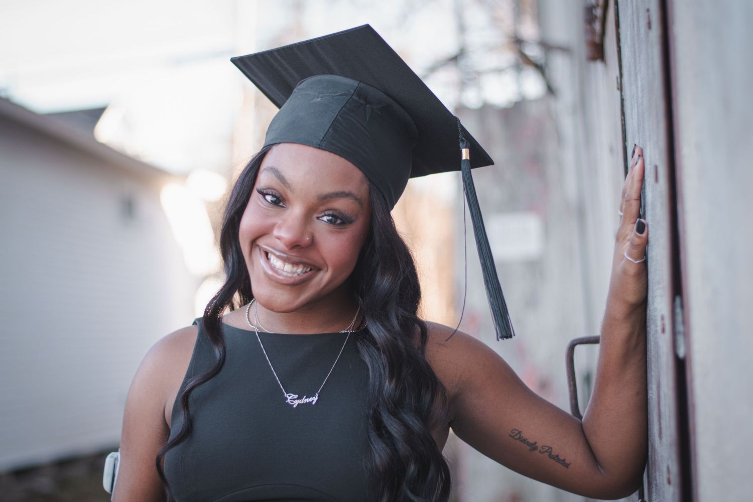 Smiling graduate woman wearing black cap and sleeveless dress, leaning against wall with tattoo visible.