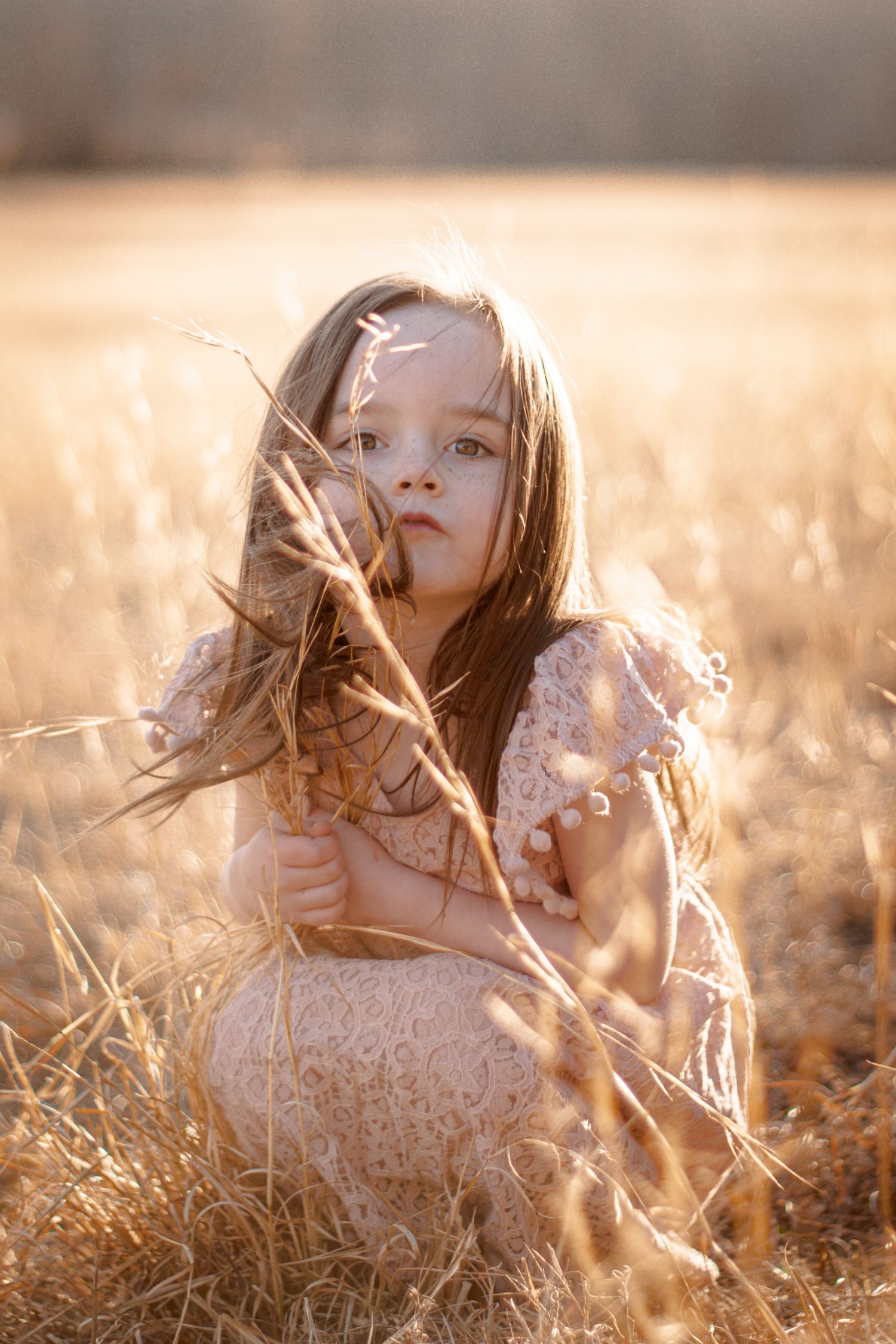 Young girl in lace dress sitting in golden field during sunset holding dried grass.