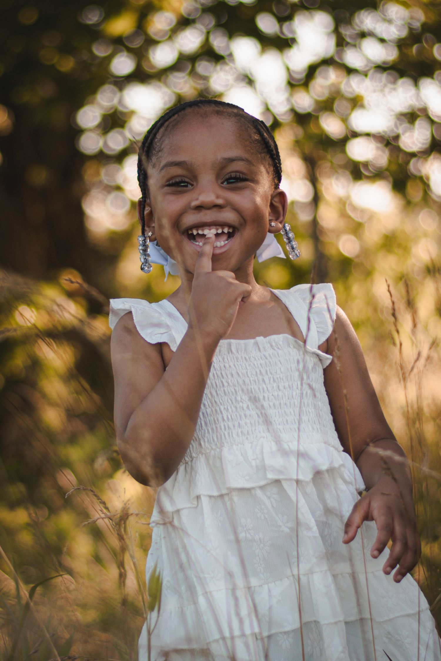 A joyful young girl in a white dress laughs outdoors, pointing to her smile in a sunlit field.
