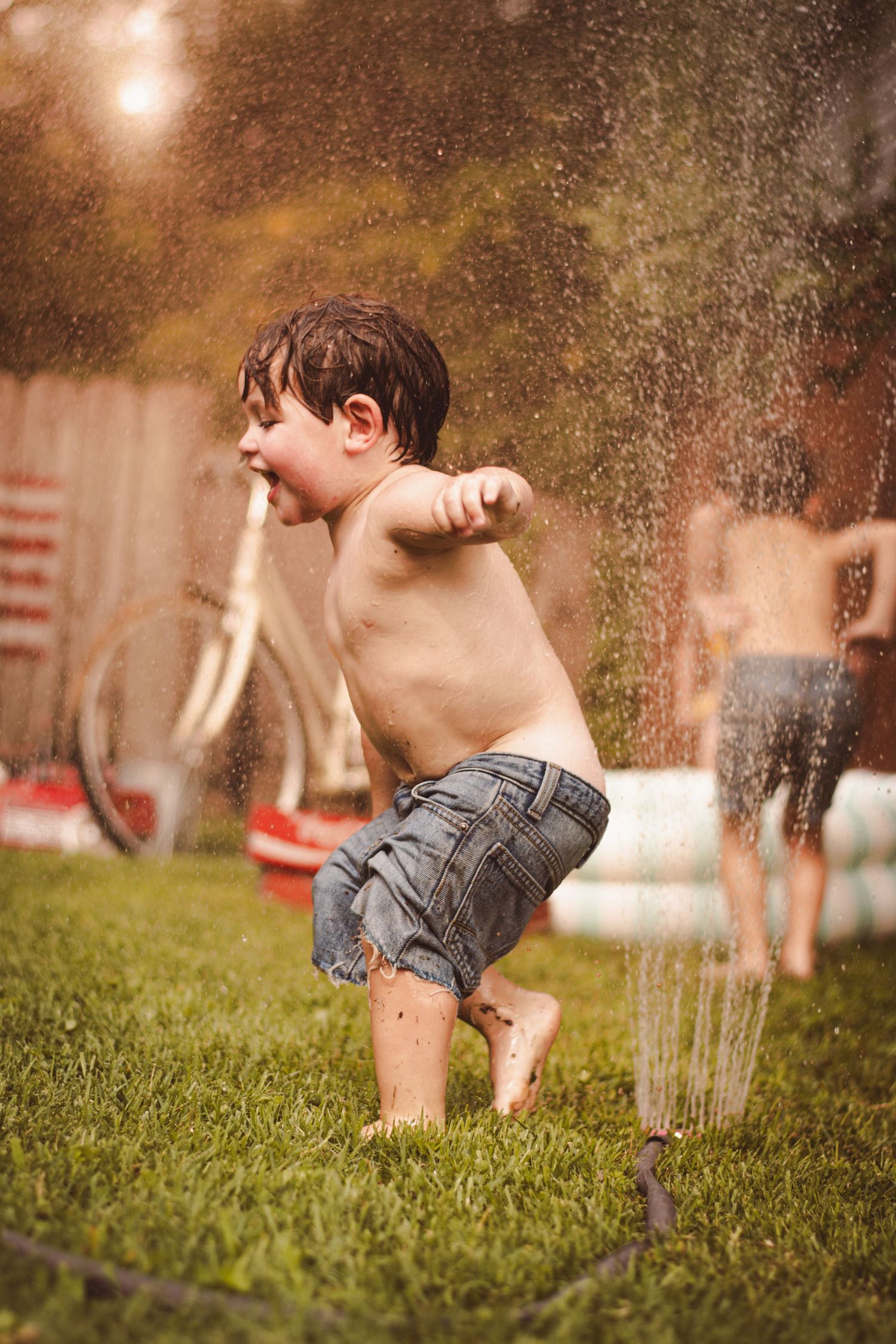 Young boy playing in backyard sprinkler on green grass during summer, wearing denim shorts, laughing with joy. Lifestyle Photography
