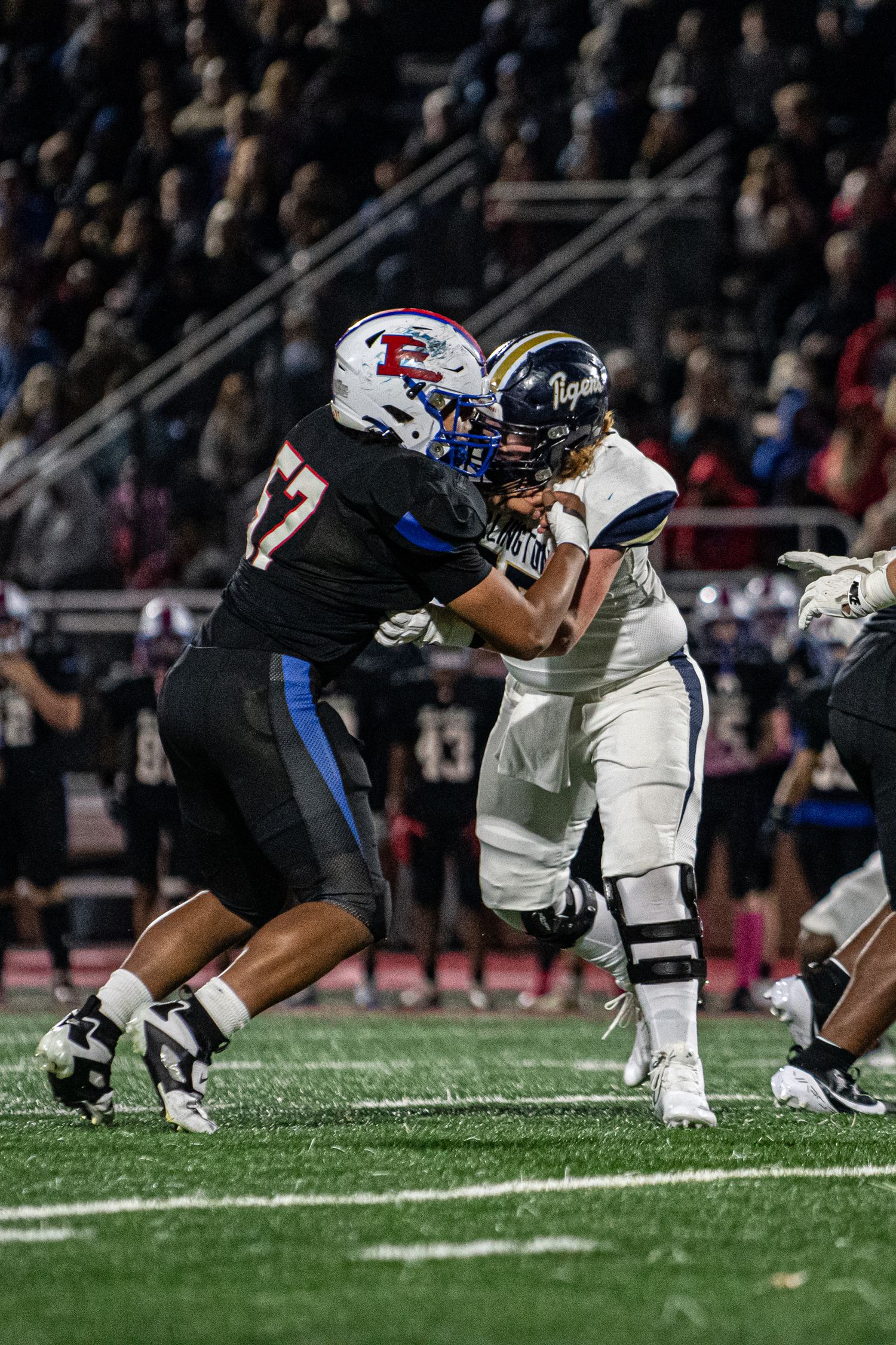 Two football players collide during a night game, with one player in black jersey number 57 blocking an opponent in white.