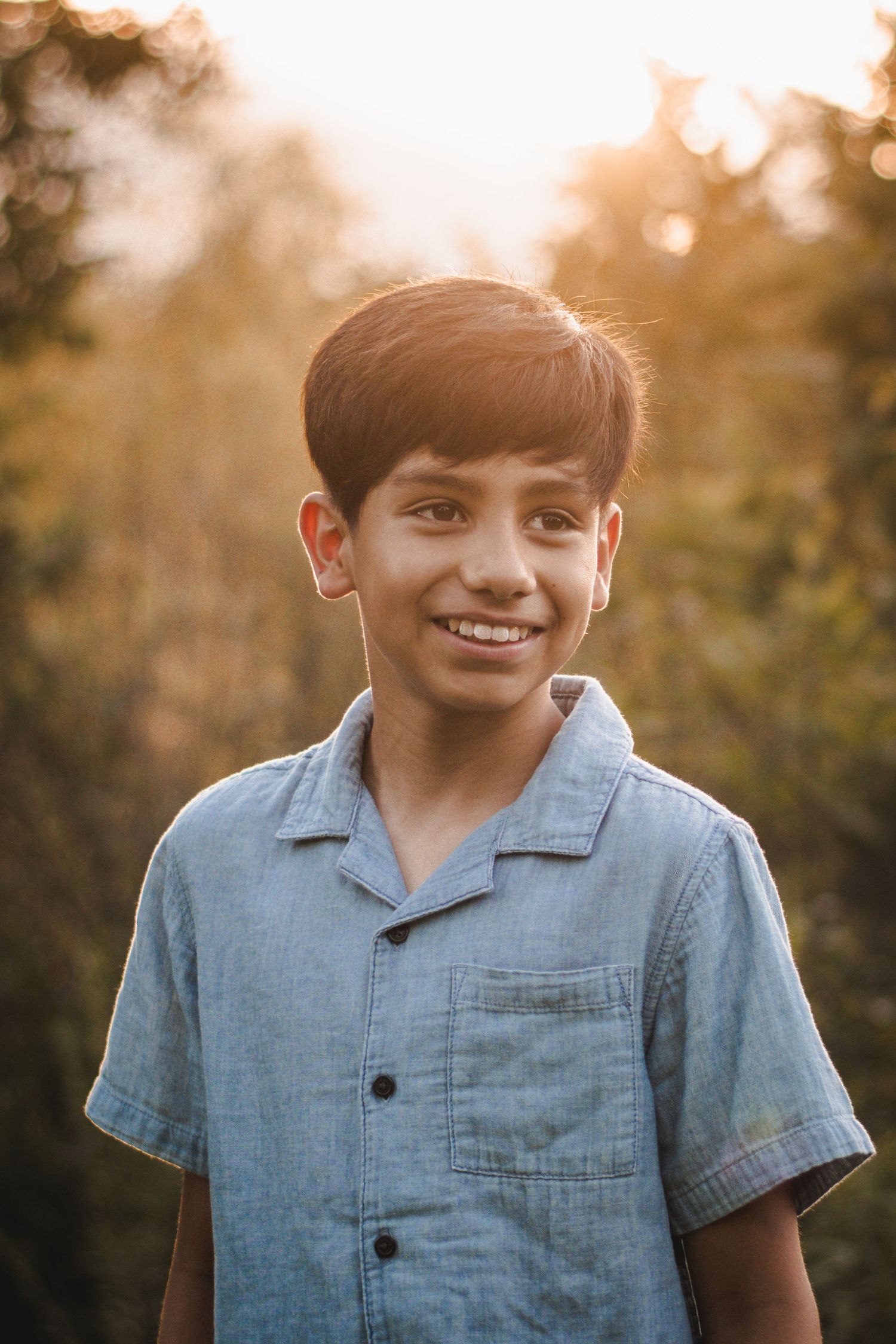 A smiling young boy in a blue chambray shirt poses outdoors with warm golden sunset light filtering through trees.