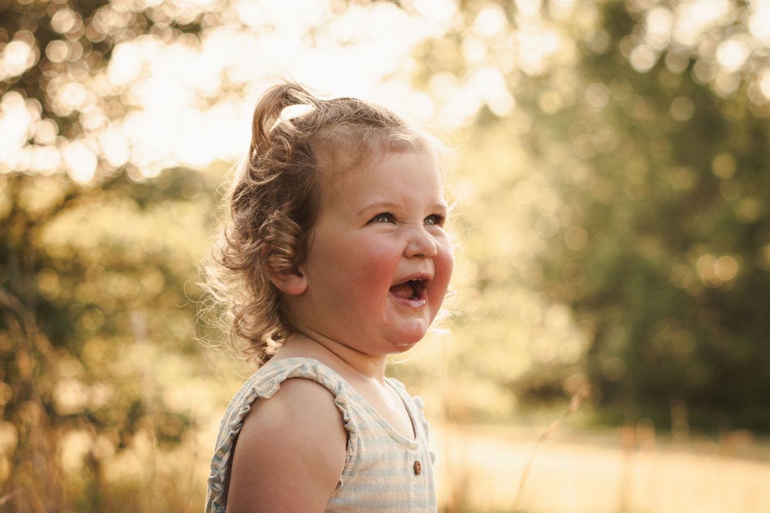 Adorable toddler girl with curly hair laughing joyfully outdoors in warm golden sunlight.