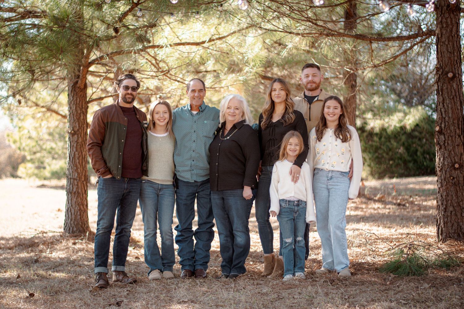 Large family of eight posing together outdoors among pine trees in casual autumn attire.