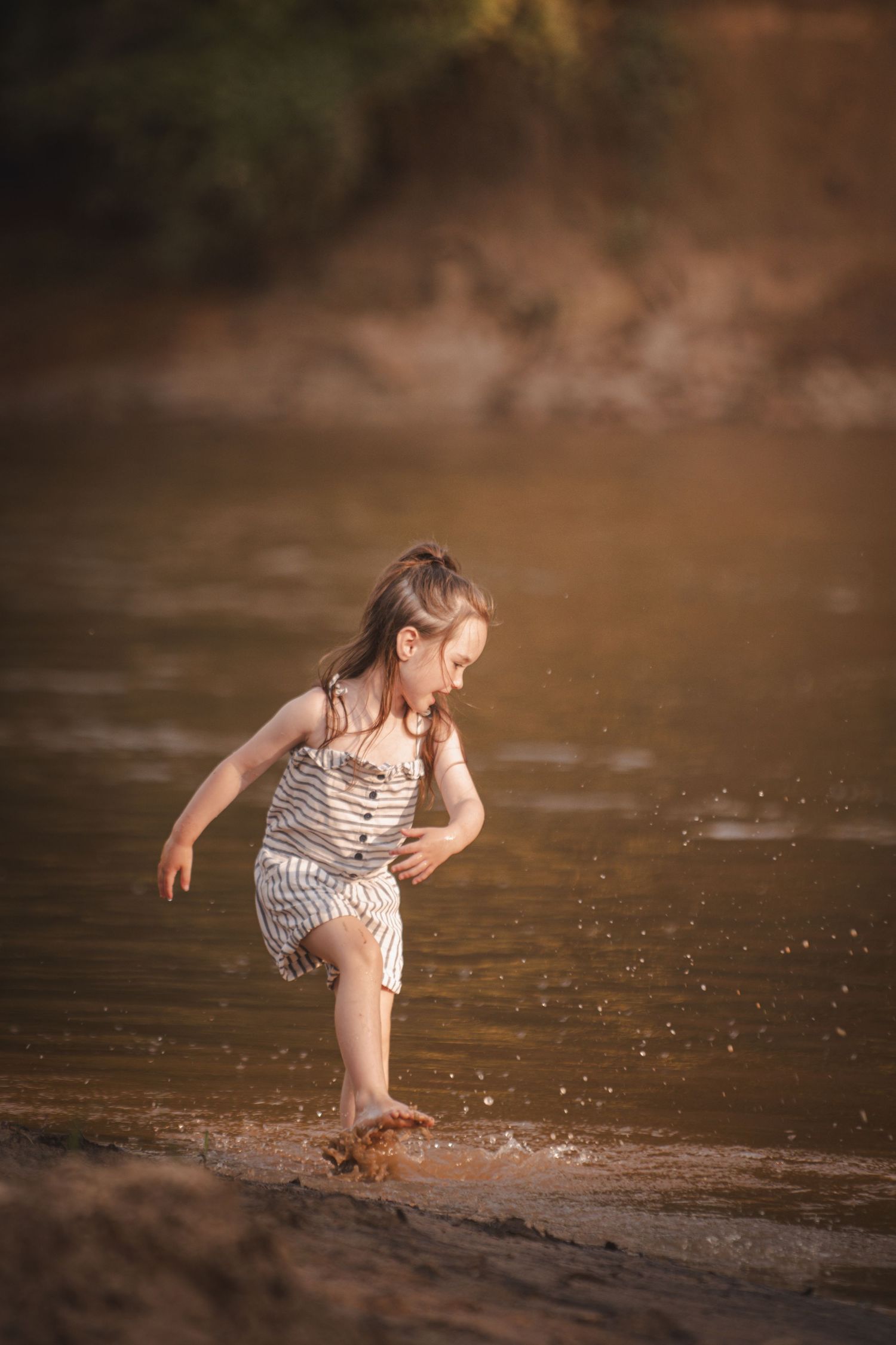 Young girl in striped romper splashing barefoot in shallow river water during golden hour.