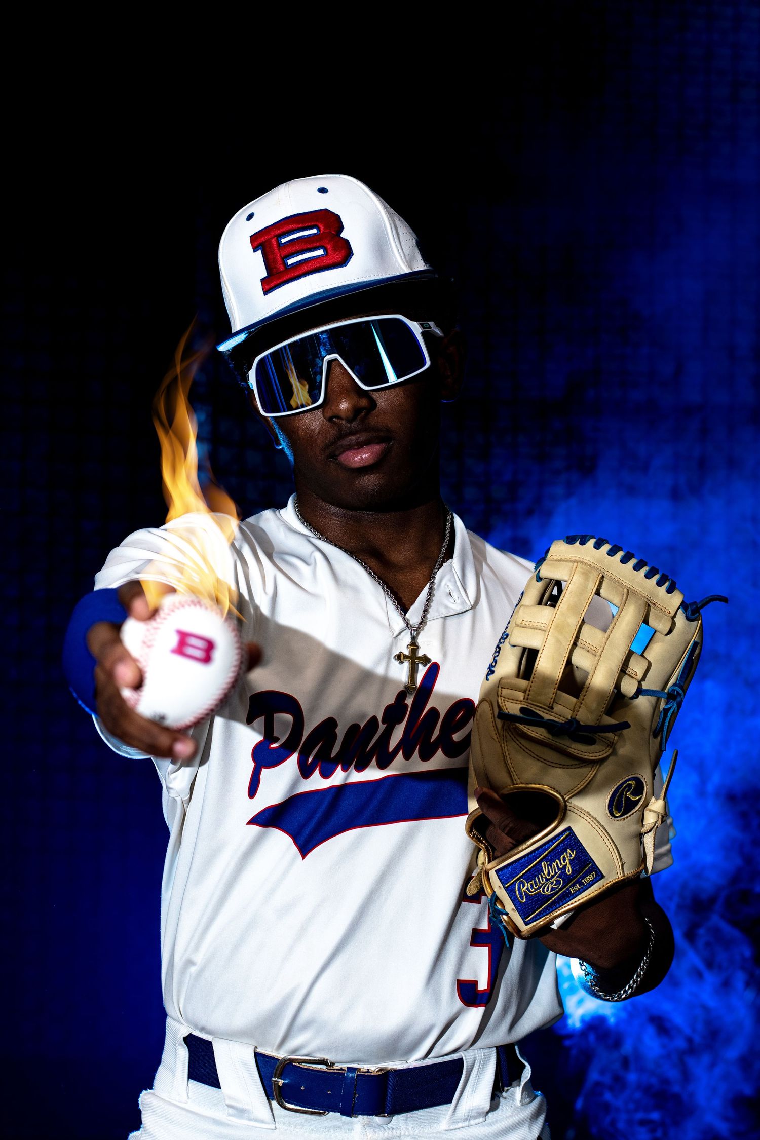 Baseball player holds a flaming ball toward camera, wearing Rawlings glove and blue shield sunglasses. Media day photoshoot with flames