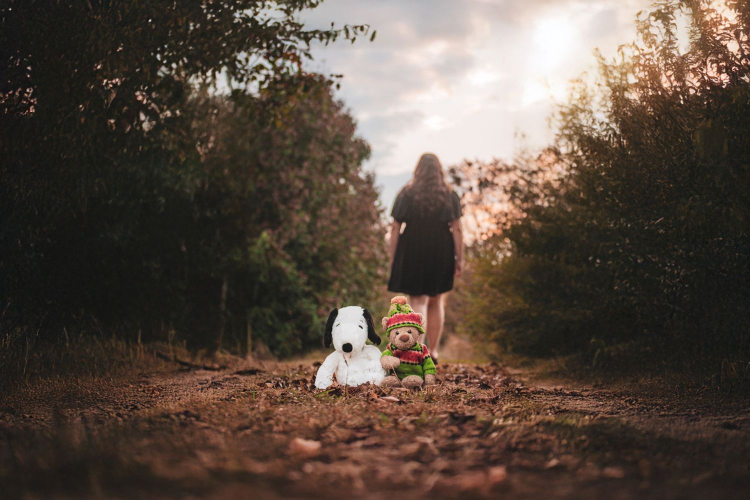 Girl in black dress walks away on dirt path leaving stuffed animals behind.