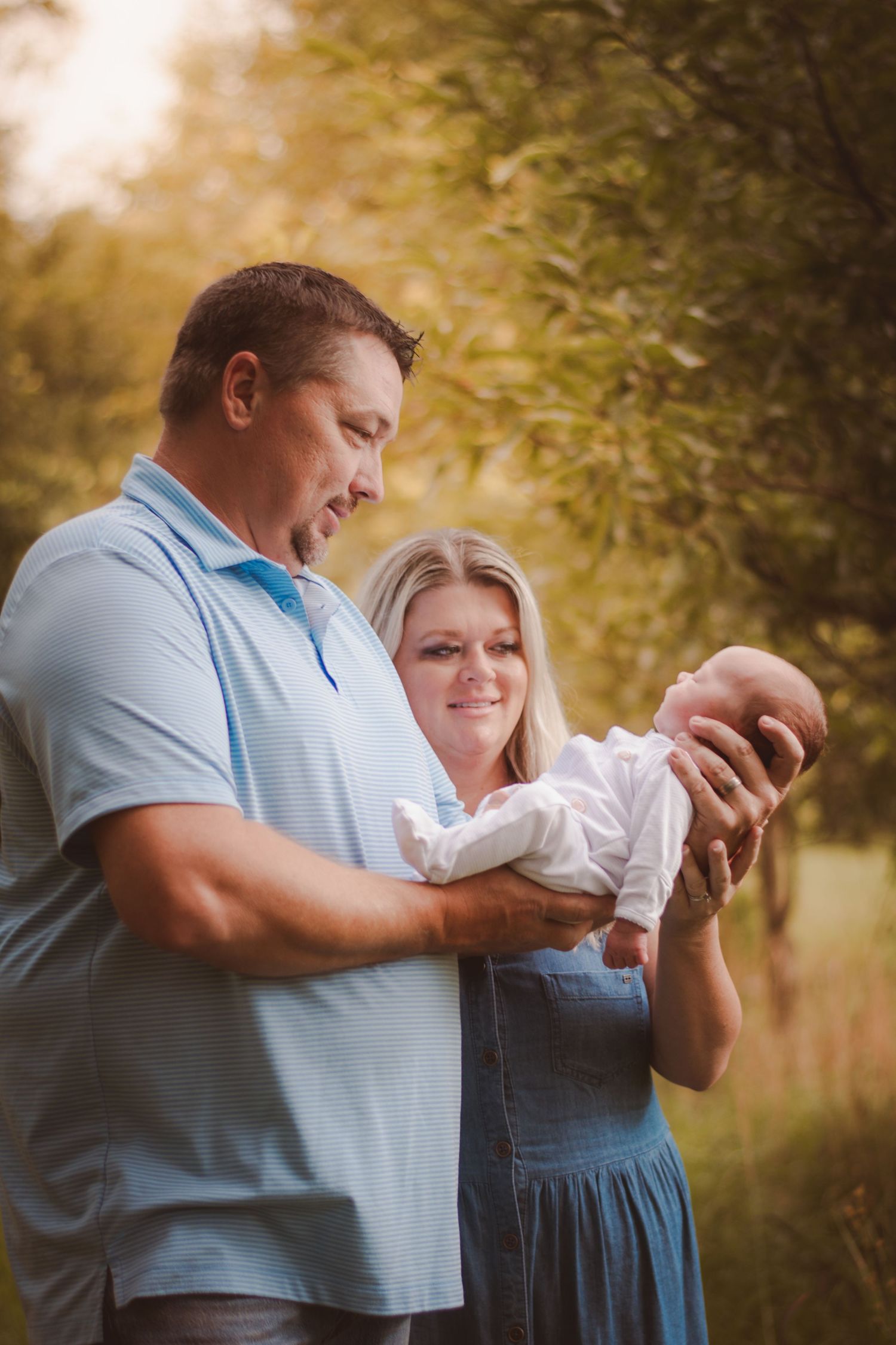 A couple holds their newborn baby outdoors in golden sunlight, surrounded by lush green trees.