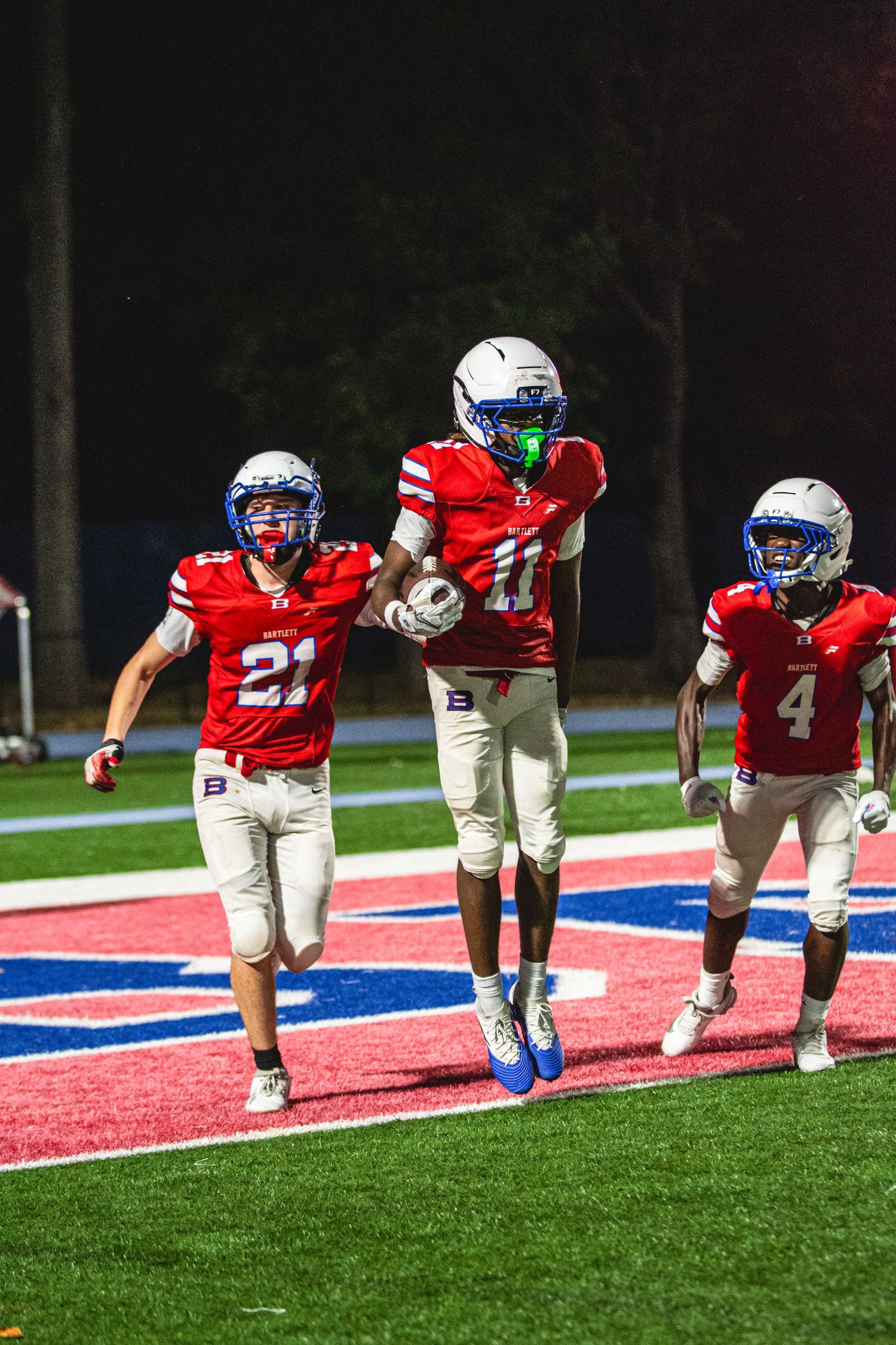 Three football players in red uniforms and white helmets celebrate on the field during a night game.