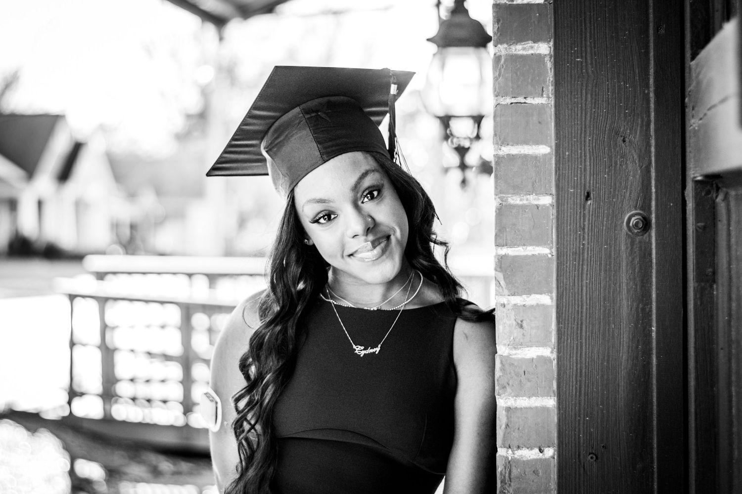 Smiling graduate in cap and black dress in black and white portrait photo.