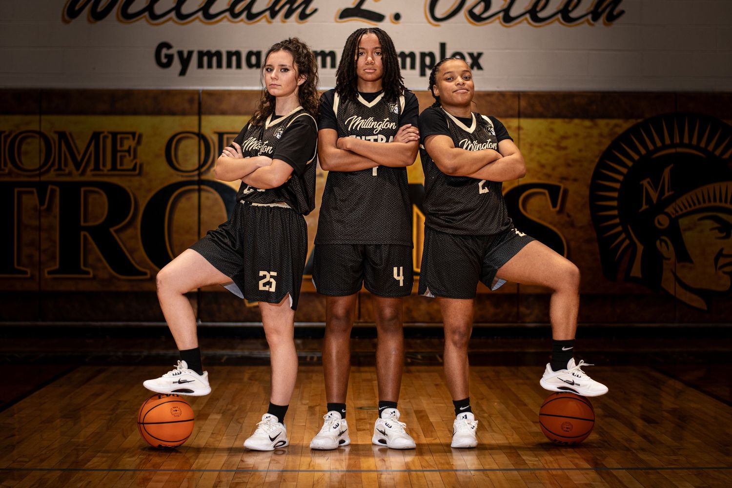 Three Millington Trojans basketball players posing confidently in a school gymnasium.
