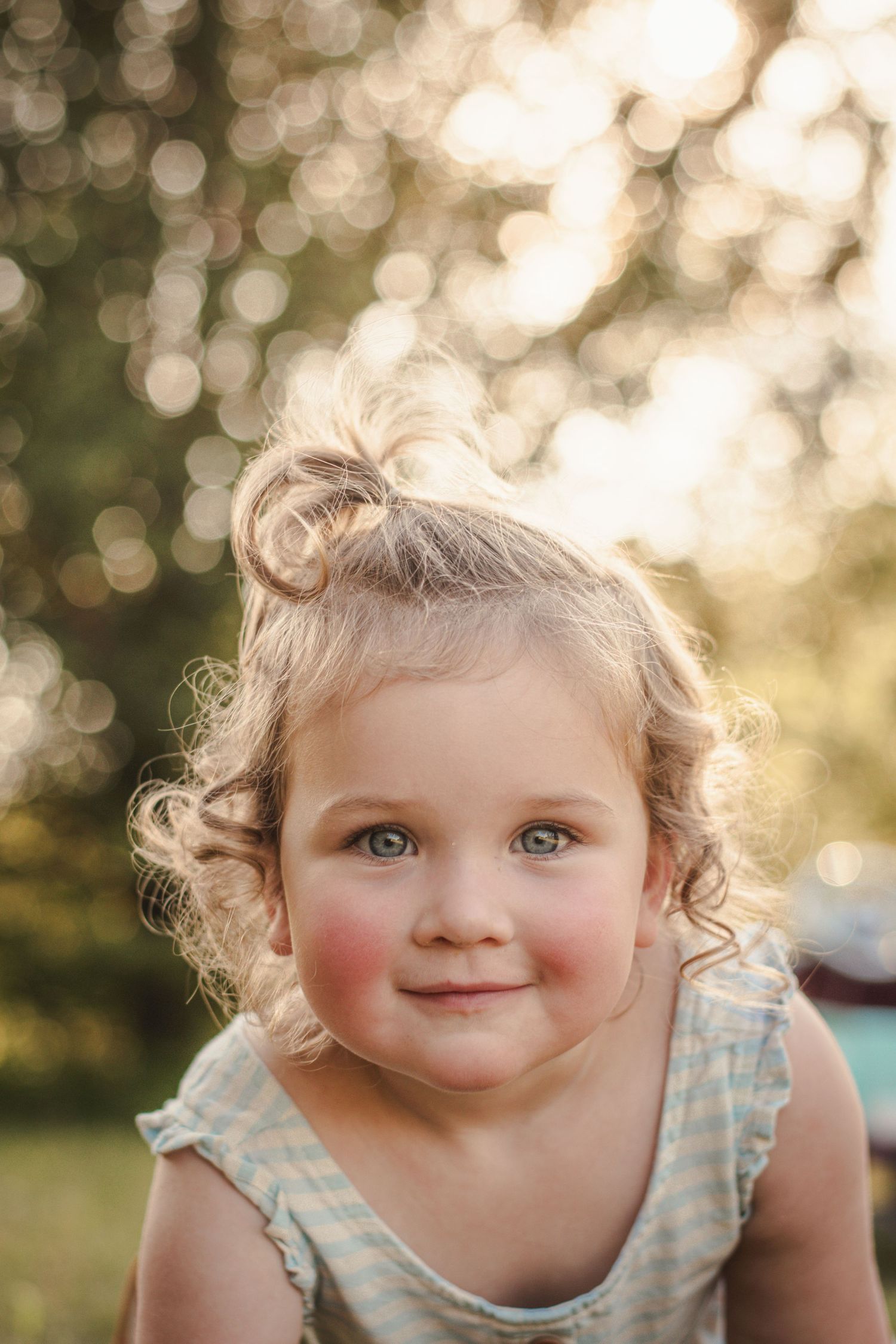Cute toddler girl with curly blonde hair and blue eyes smiling in warm golden sunlight.