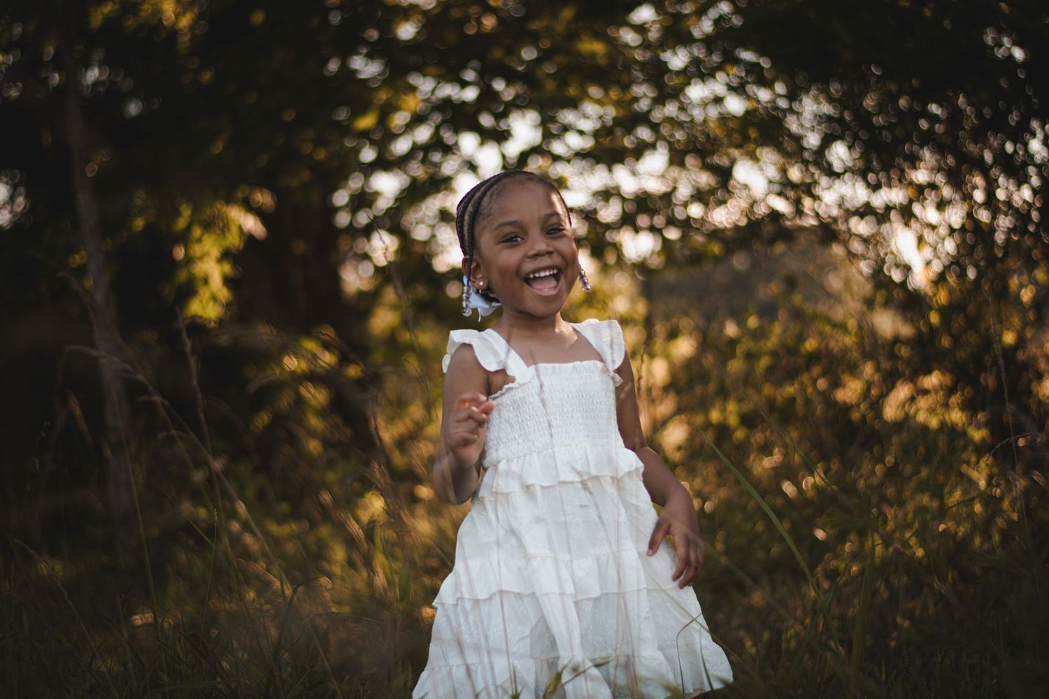 A joyful young girl in a white dress laughs while playing outdoors in golden sunlight among lush green trees.