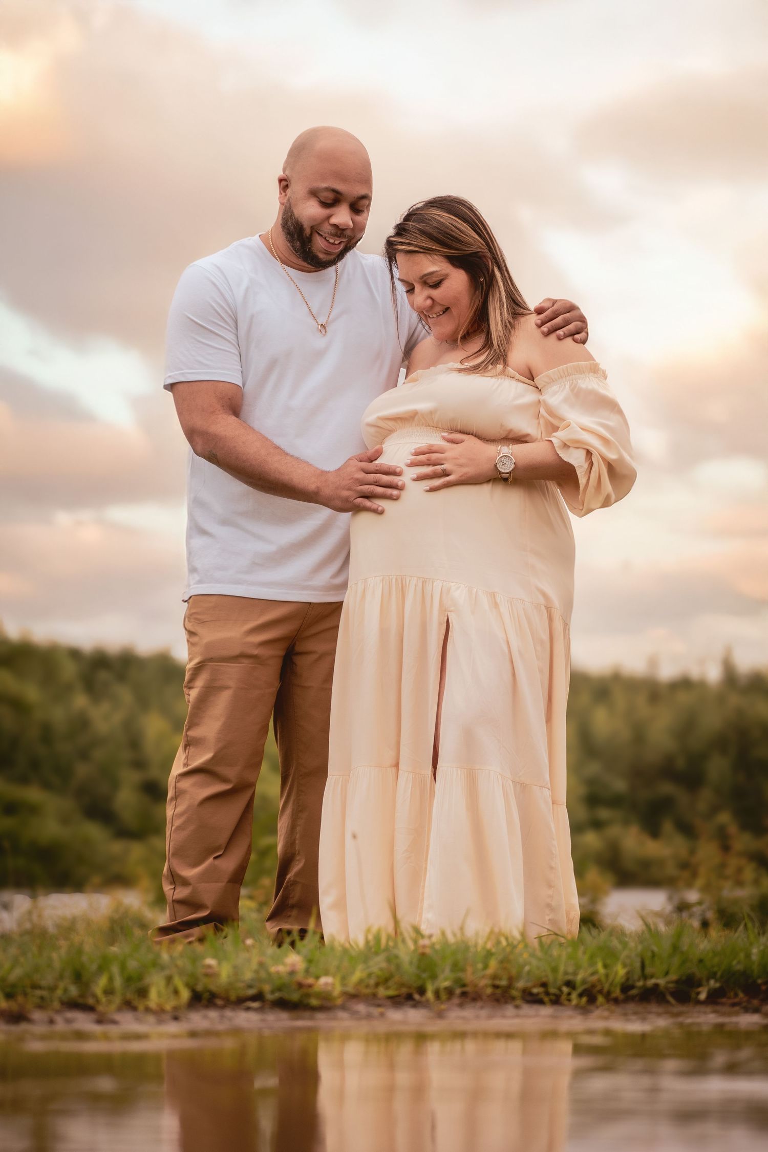 Couple sharing a tender maternity photo moment by a reflective pond at sunset.