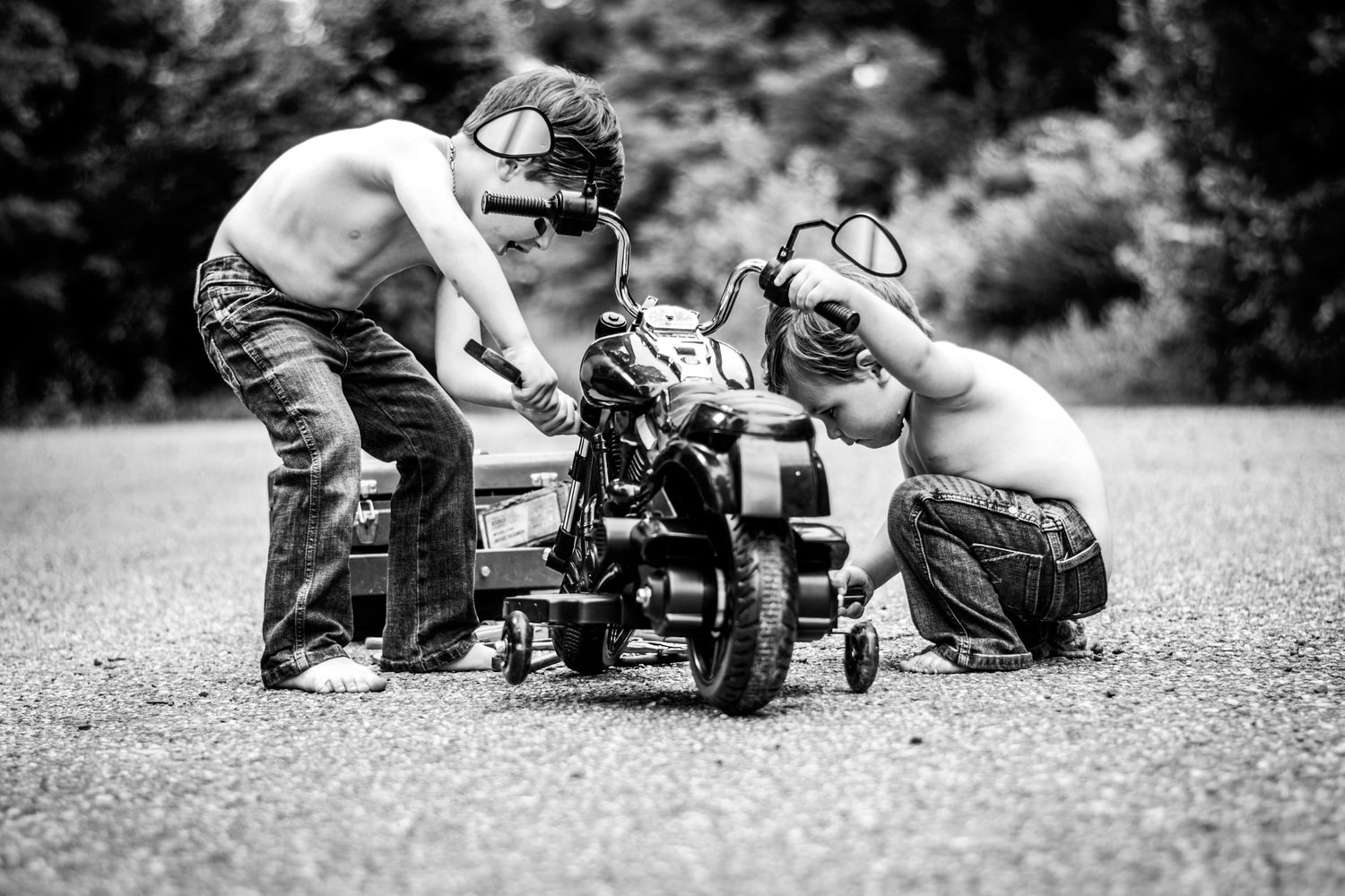 Two shirtless boys wearing goggles work on a small motorcycle on pavement in a black and white photo.