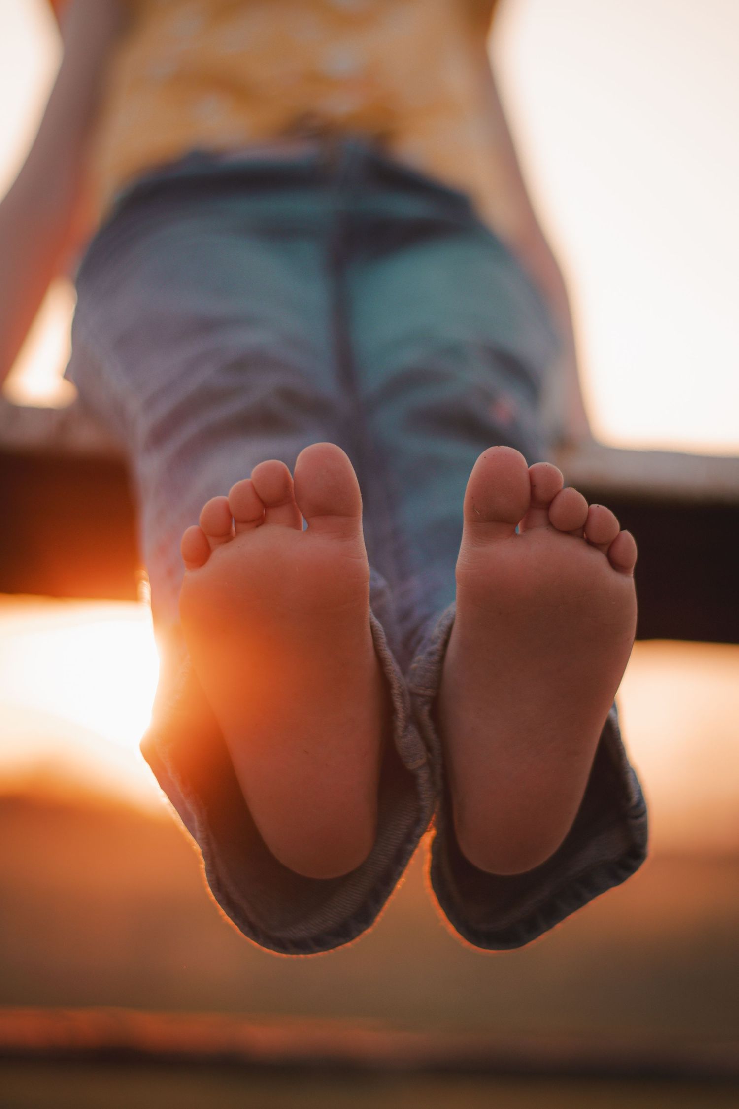Child's bare feet dangling at sunset, wearing blue jeans, warm golden backlight creating a serene summer evening scene.