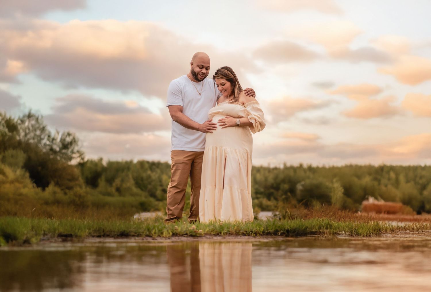 Expecting couple stands by reflective water at sunset, woman in flowing cream maternity gown, man in white shirt.