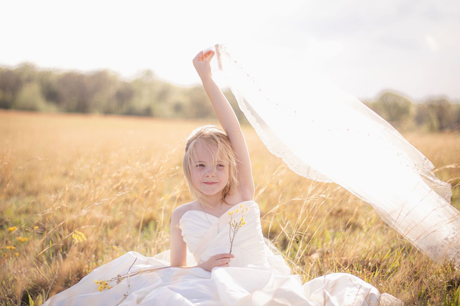Young girl in a white flower girl dress sits in a sunny golden field holding up a veil.