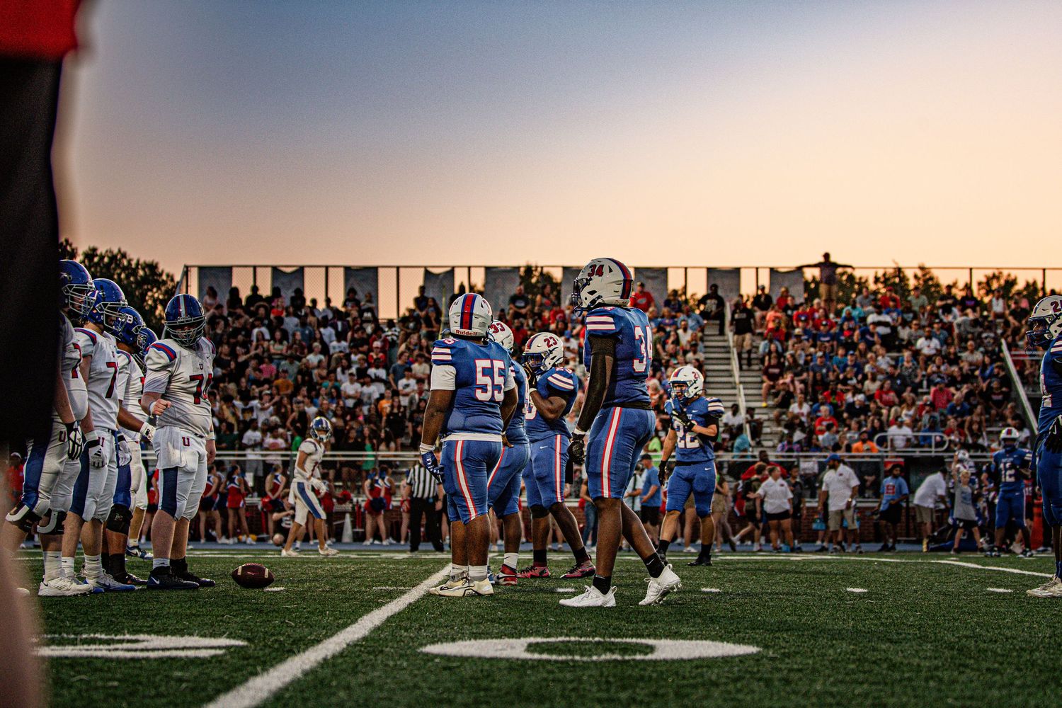 High school football players on field at sunset with packed stadium crowd in background.