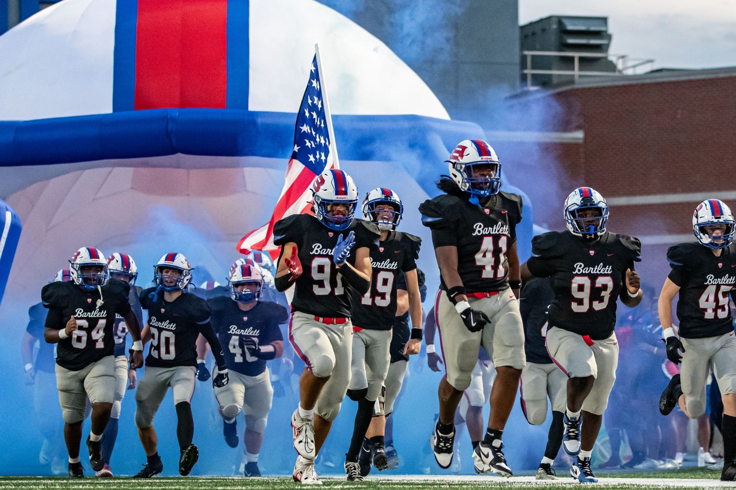 High school football team running onto field carrying American flag.