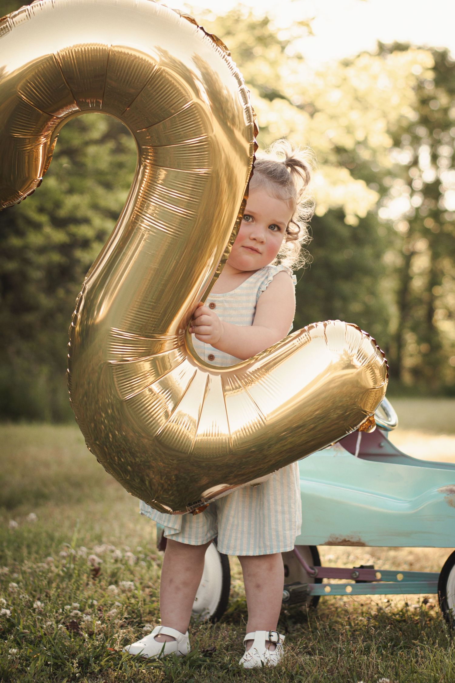 Toddler girl in white dress hugging a large gold number 2 balloon outdoors at a 2nd birthday celebration.
