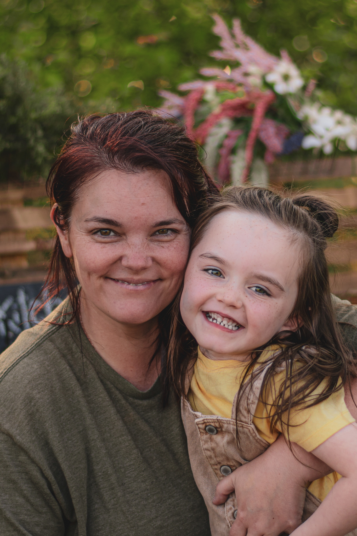 A smiling woman with red hair hugs a cheerful young girl in a yellow shirt outdoors near colorful flowers.