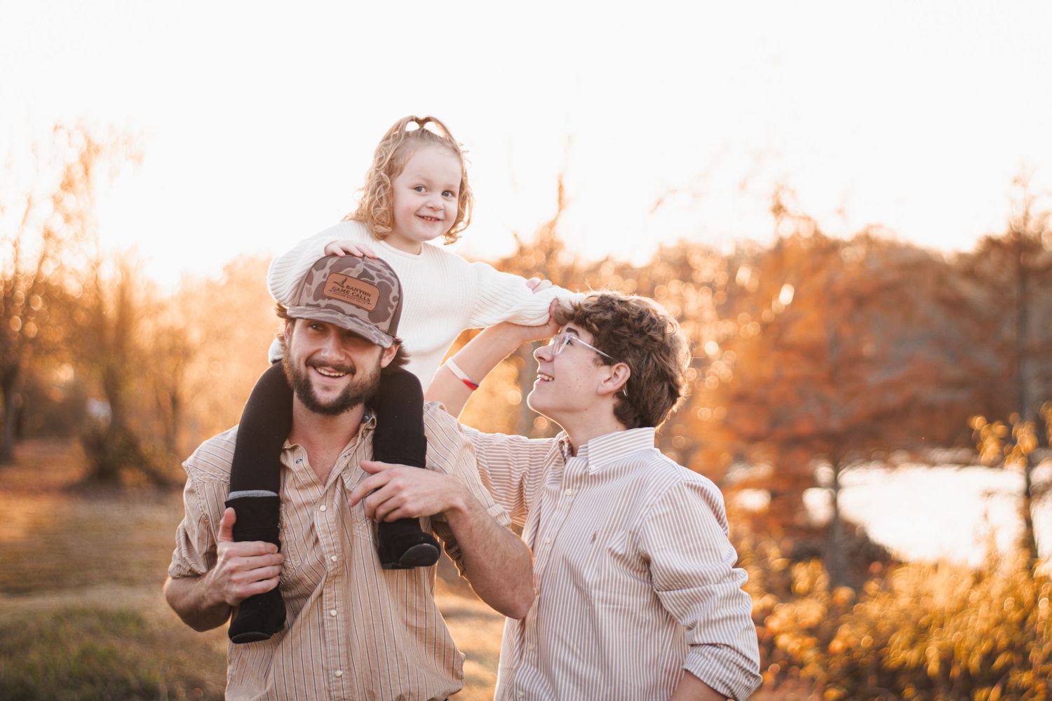 Two dads enjoying a sunny autumn day outdoors with their young daughter sitting on shoulders.