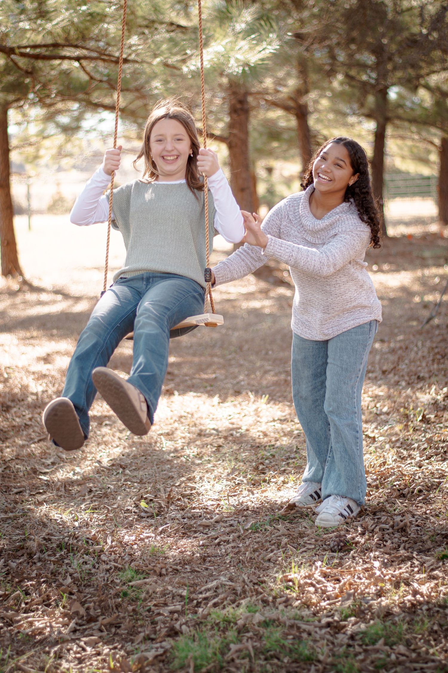 Two girls laughing outdoors, one on a rope swing as the other pushes her in a sunny pine forest setting.
