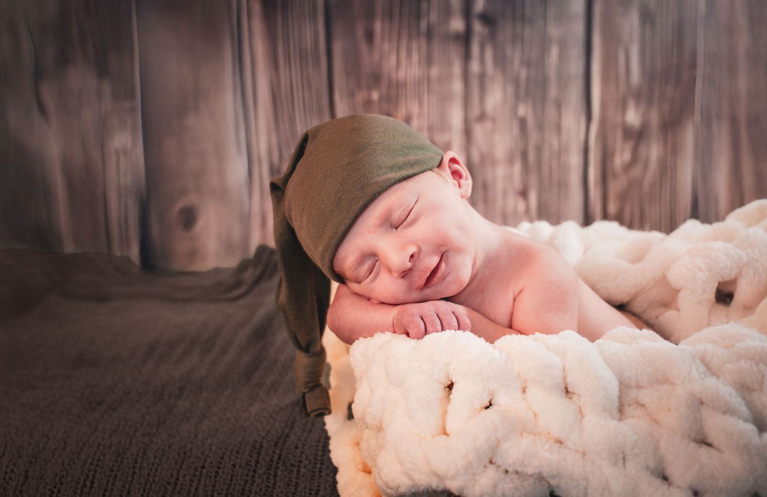Peaceful smiling newborn baby sleeping in a bowl wearing a green knit hat on blanket.