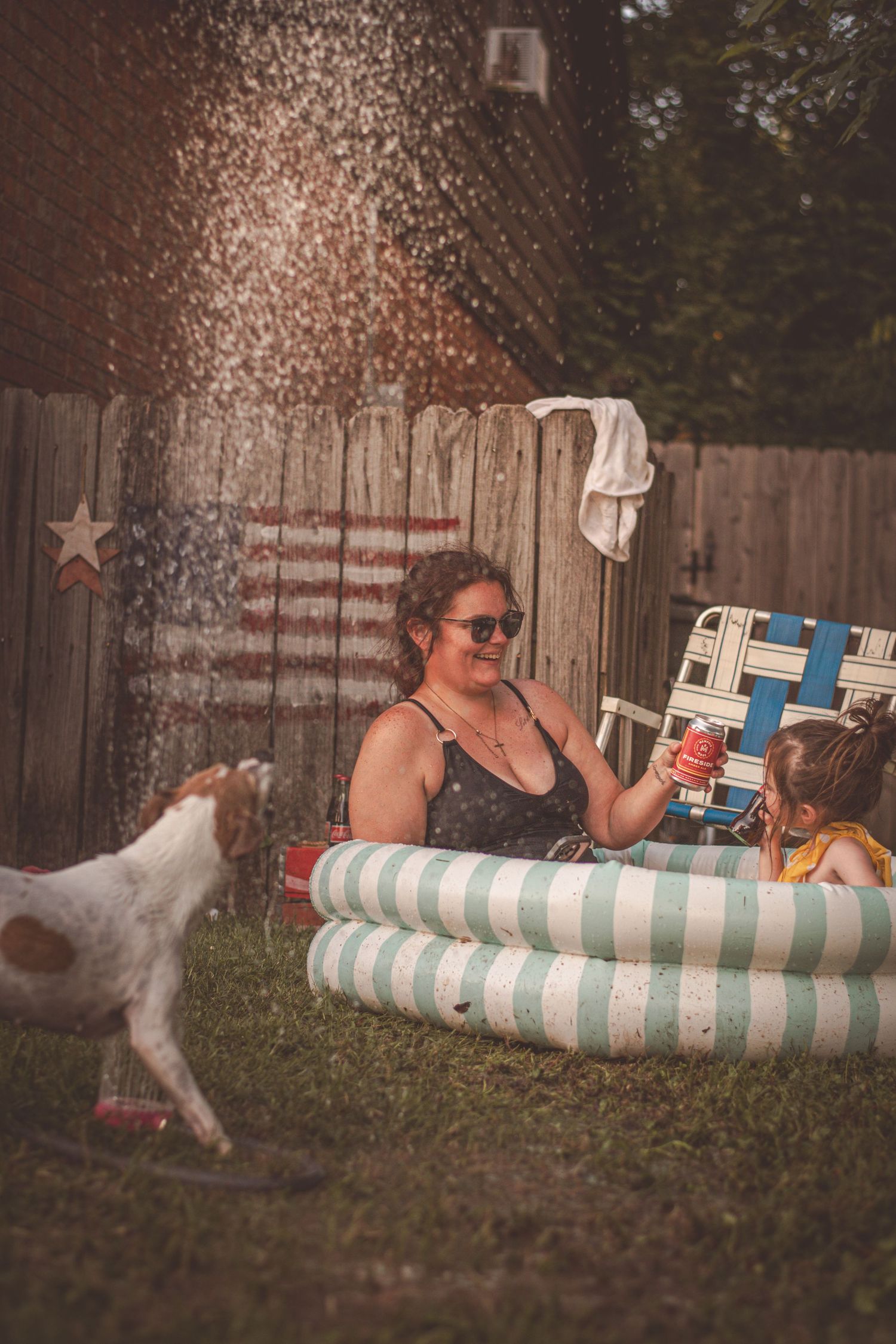 Woman and child playing in inflatable pool in backyard with dog and American flag on fence.