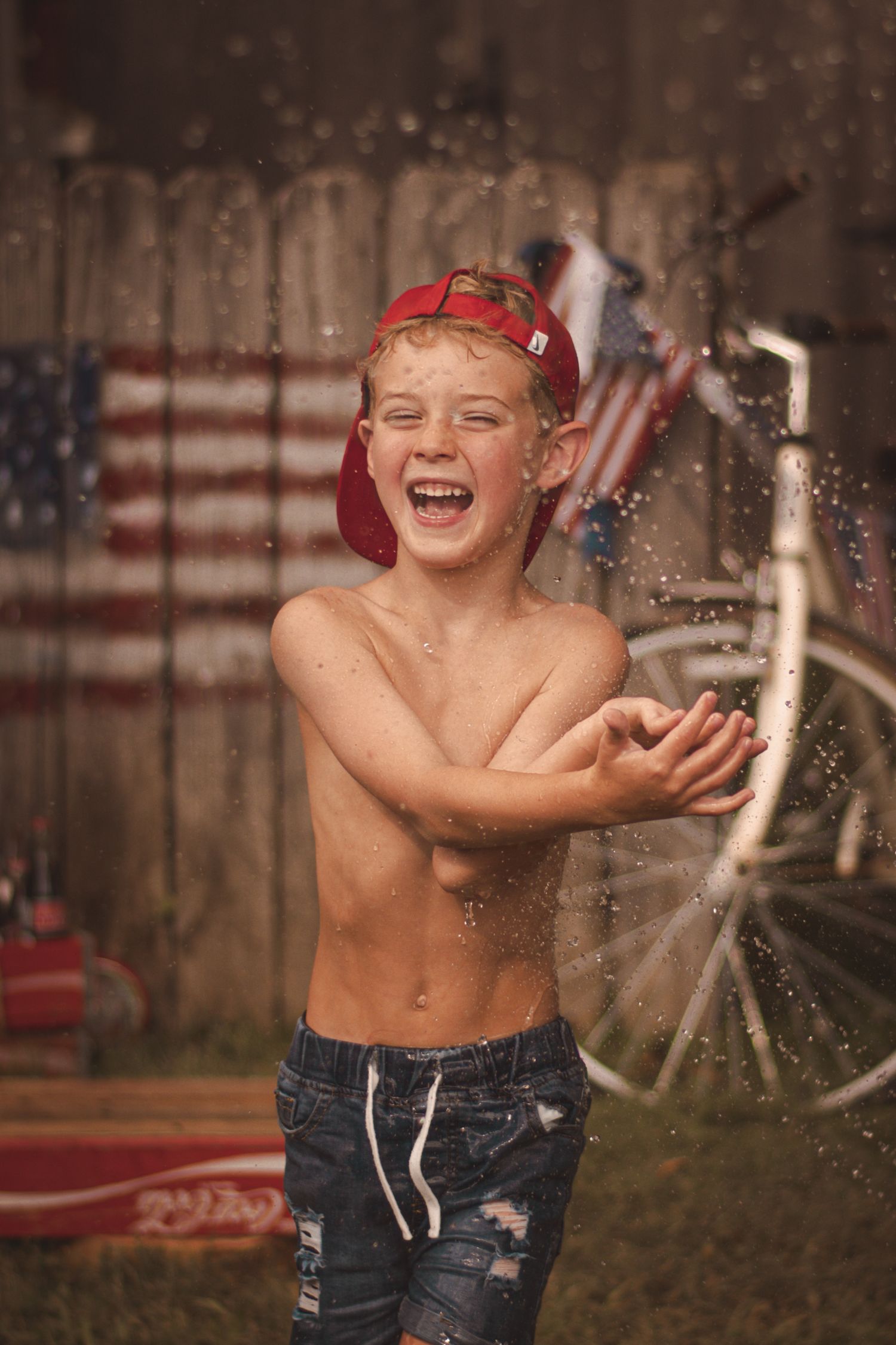 A laughing shirtless boy in a red cap plays in water sprinkles outdoors with an American flag and bicycle in the background.