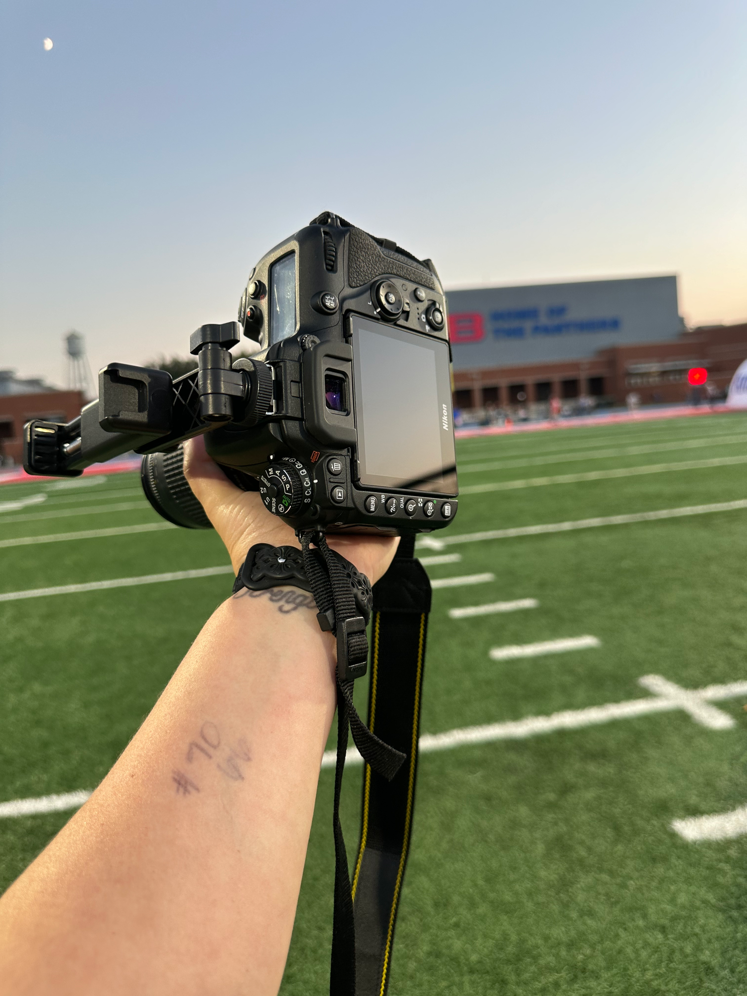 A hand holding a Nikon DSLR camera on a tripod at a football field during dusk.