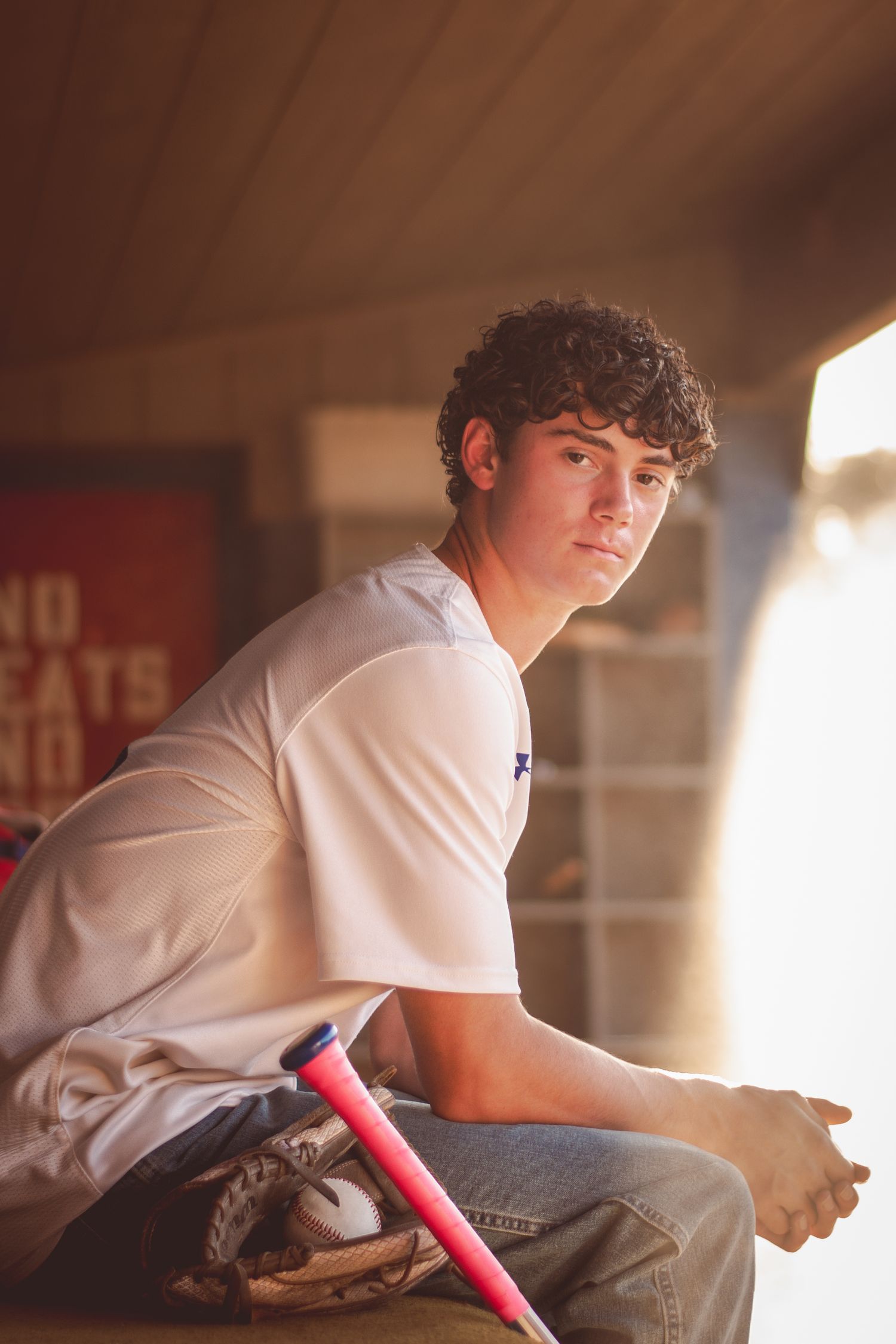 Young baseball player sitting in dugout with glove and pink bat, wearing white jersey, portrait photo.