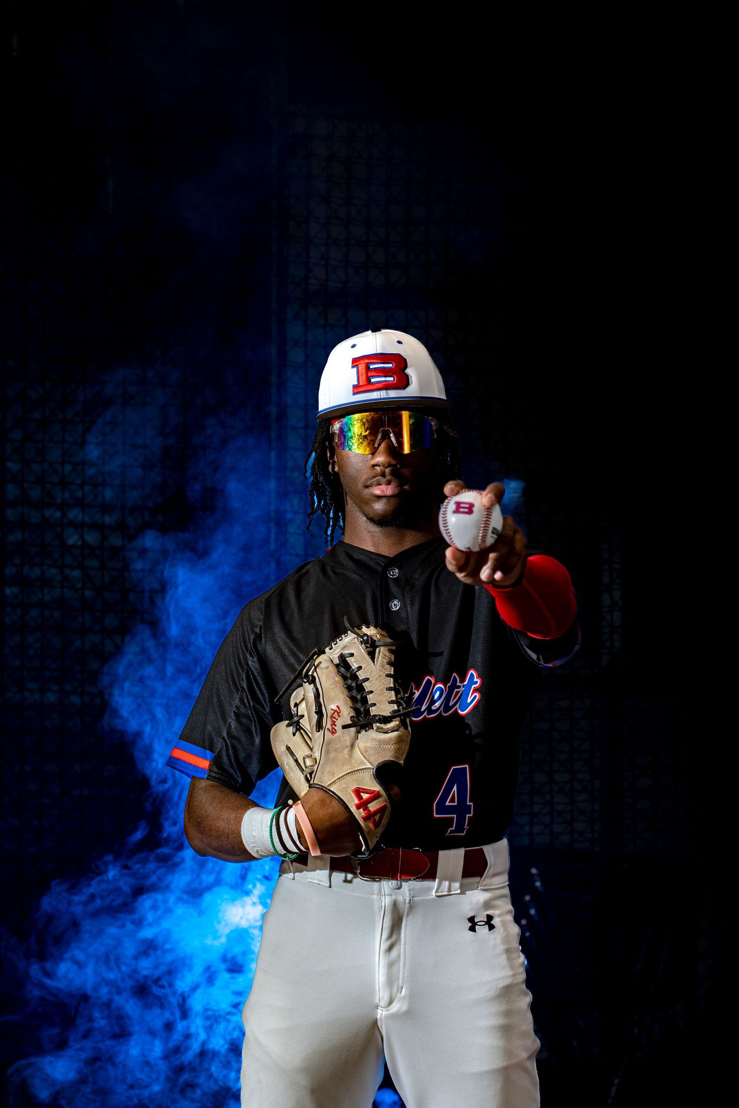 Baseball player in black uniform with glove and ball poses dramatically against dark background with blue smoke effects.