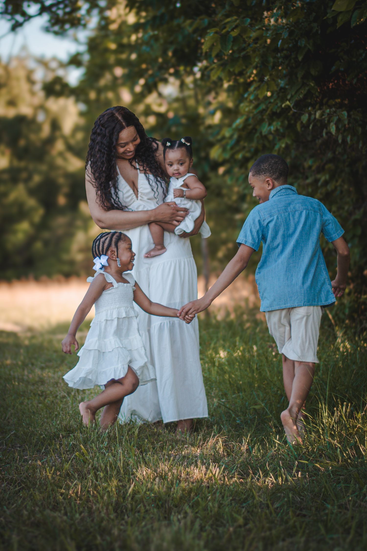 A mother in a white dress holds her baby while her two children play outdoors in a sunny green park.