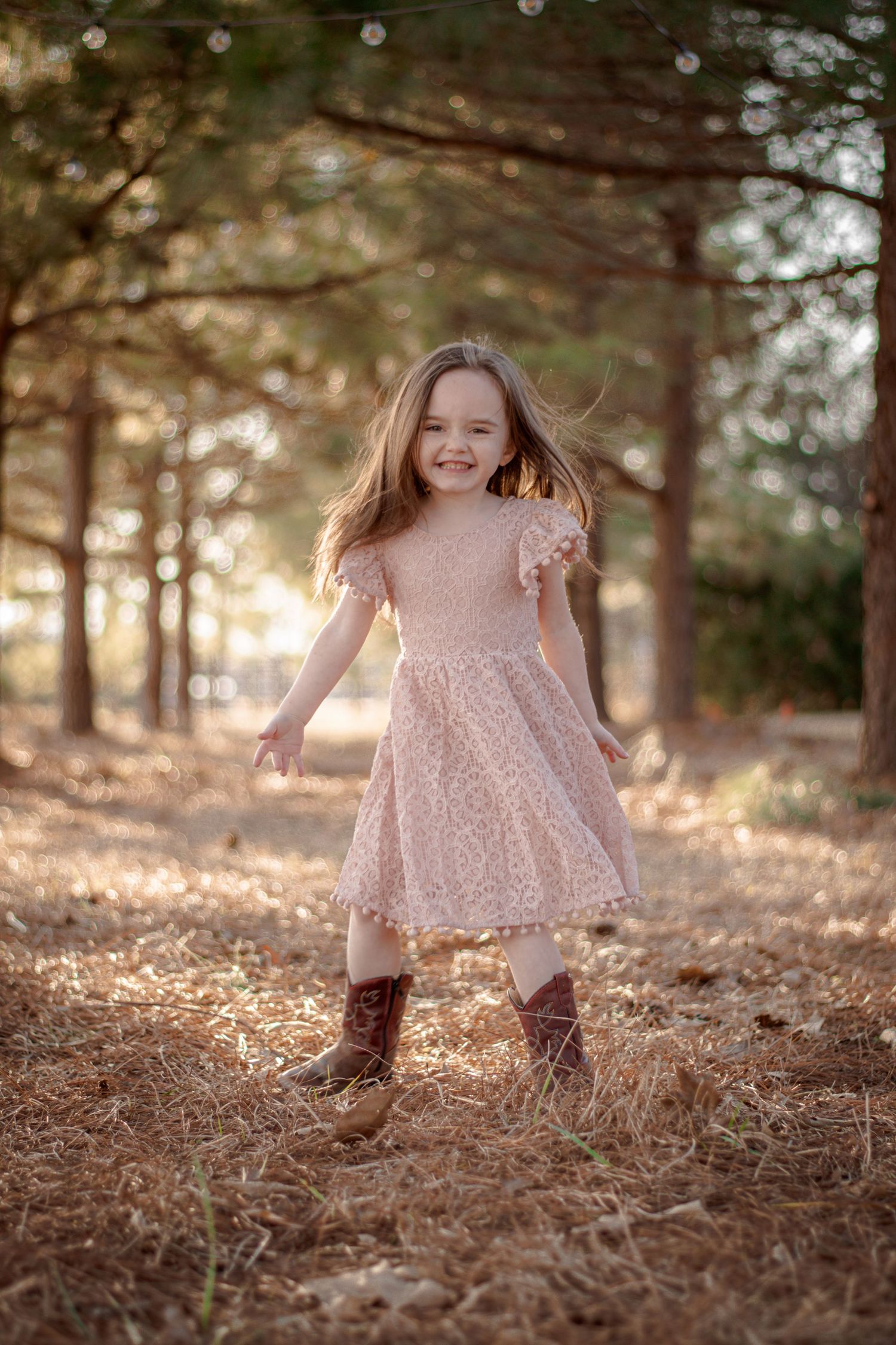 Young girl in pink lace dress and cowboy boots smiling joyfully in a sunlit pine forest path.