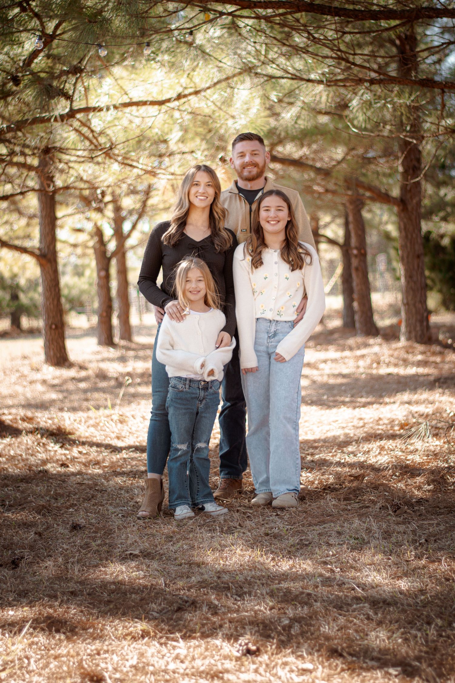 Family of four smiling together among pine trees in a sunlit forest, wearing casual jeans and sweaters.