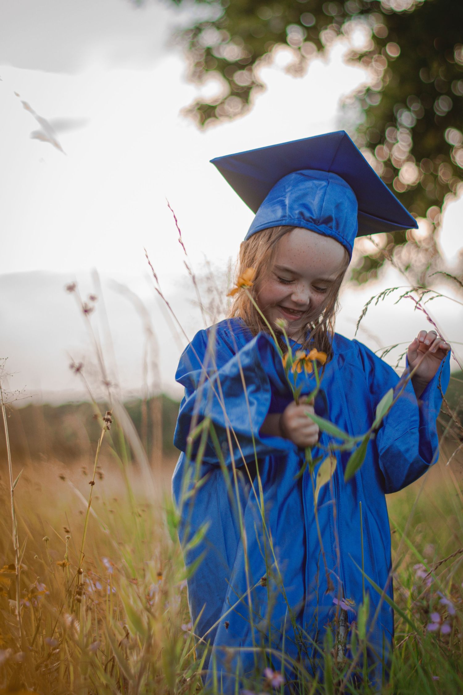 Young girl in blue graduation cap and gown smiling while holding wildflowers in a sunny meadow.