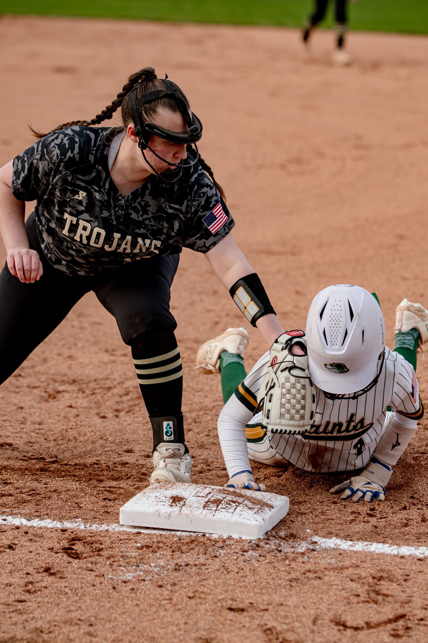 Softball player in Trojans uniform tags runner sliding into base during game.