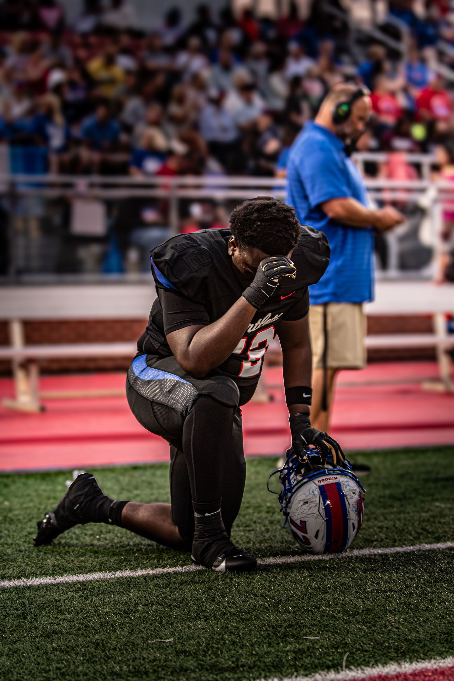 Football player kneeling in prayer on field with helmet beside him.
